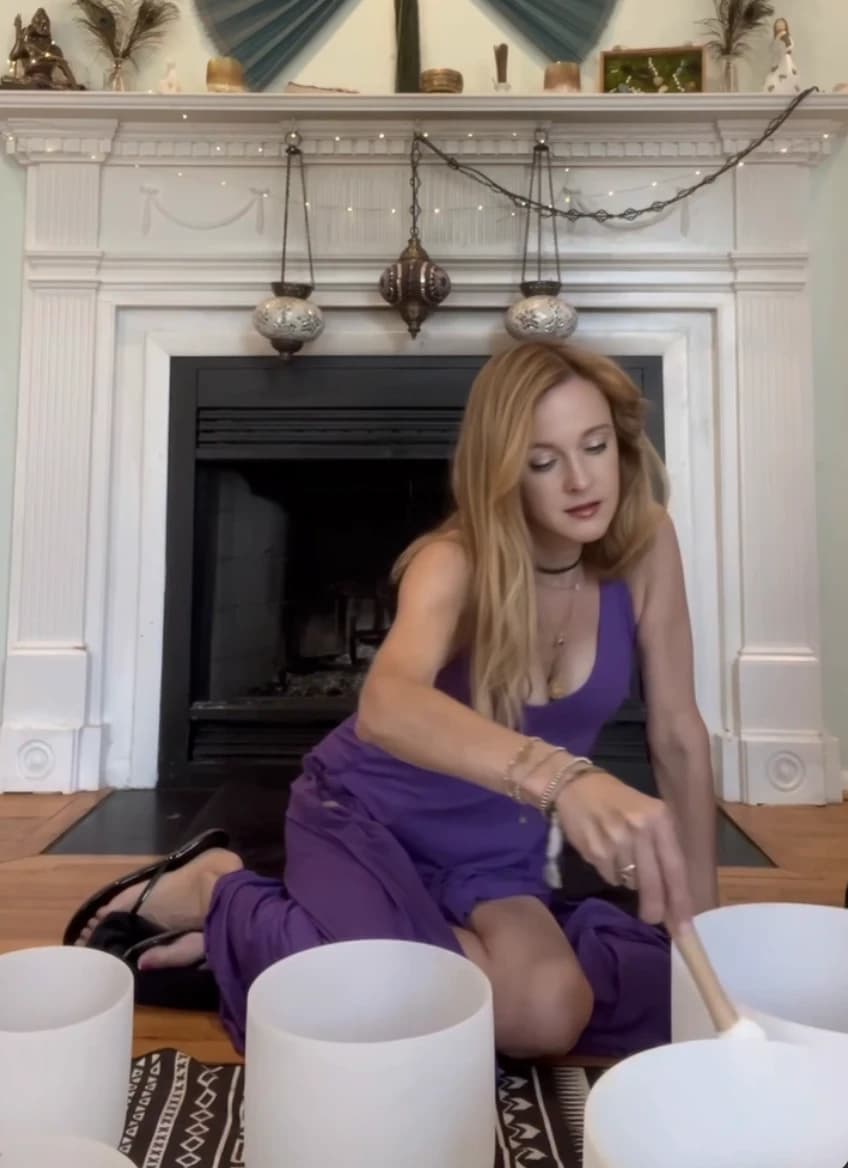 Andrea Powell playing white crystal singing bowls in front of a fireplace